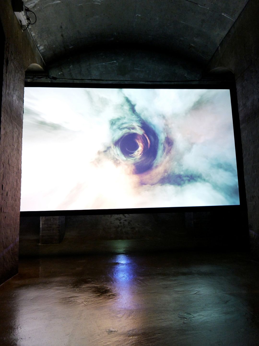 A large screen glows in the dark Copenhagen cisterns, showing a swirling eye, its light reflected on the wet stone floor.