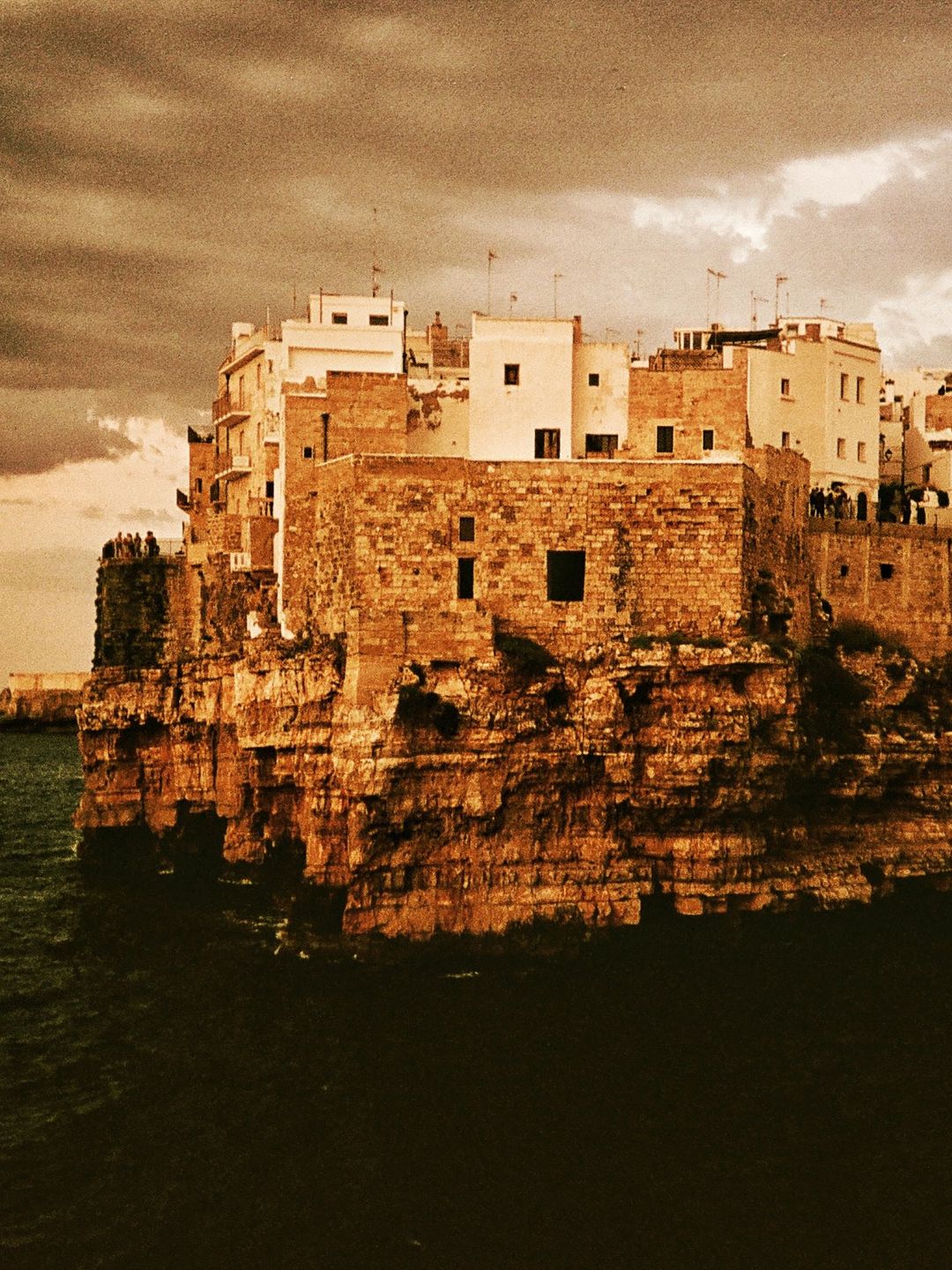 The cliff-top houses of Polignano a Mare above the sea under a stormy sky, shot on 35mm film in warm amber tones.