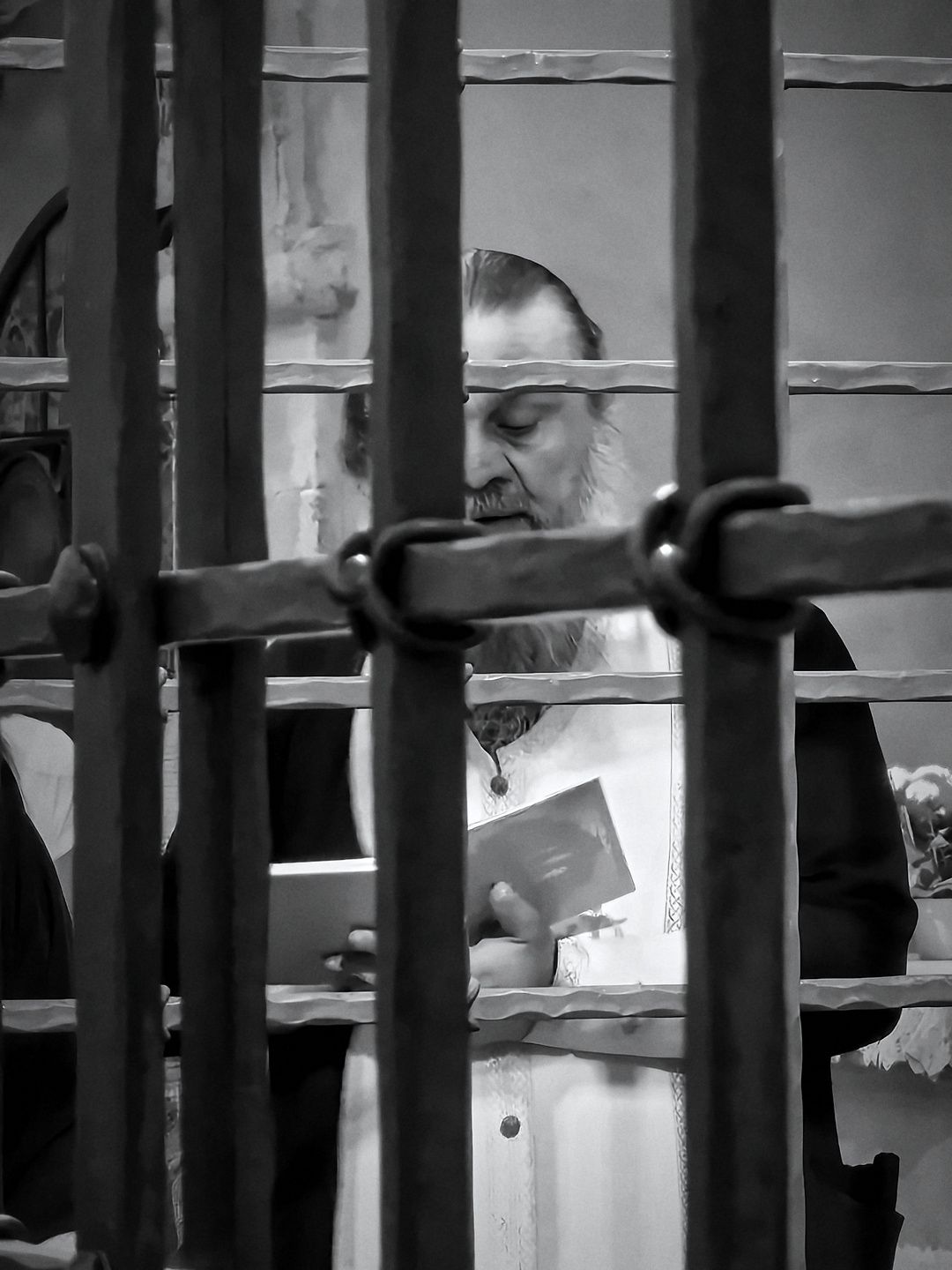 Black and white: a bearded priest in white vestments reads from a small book behind an iron grille in Bari.