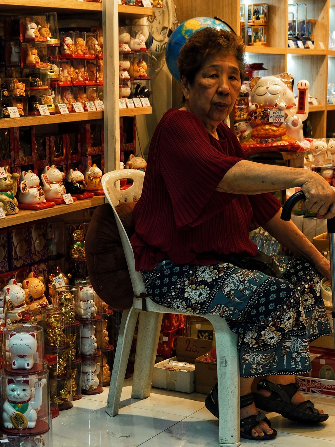 An elderly woman in a red blouse sits with a walking stick in a lucky-cat shop in Bangkok's Chinatown, surrounded by rows of maneki-neko, looking at the camera.